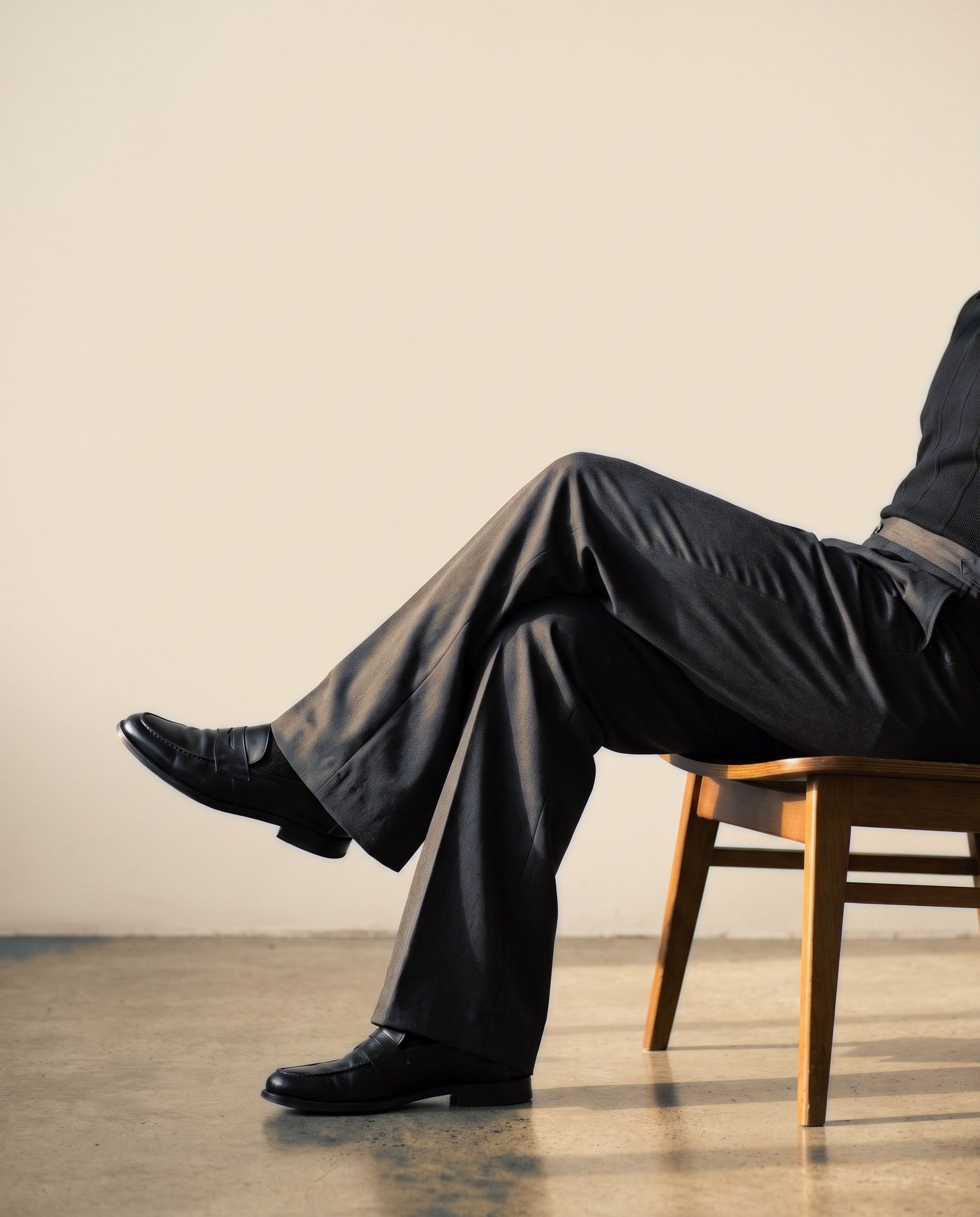 Seated model in Drip Club tailored black wool trousers and black leather loafers against a cream wall